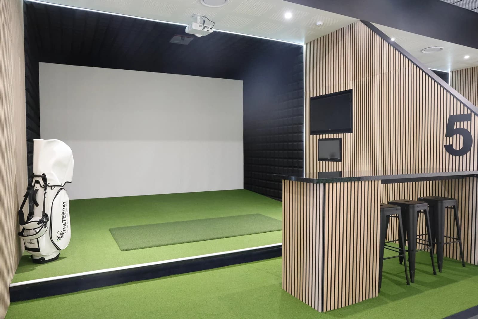 Standard golf simulator bay with a golf bag, green hitting mat and timber seating at The Tee Bay